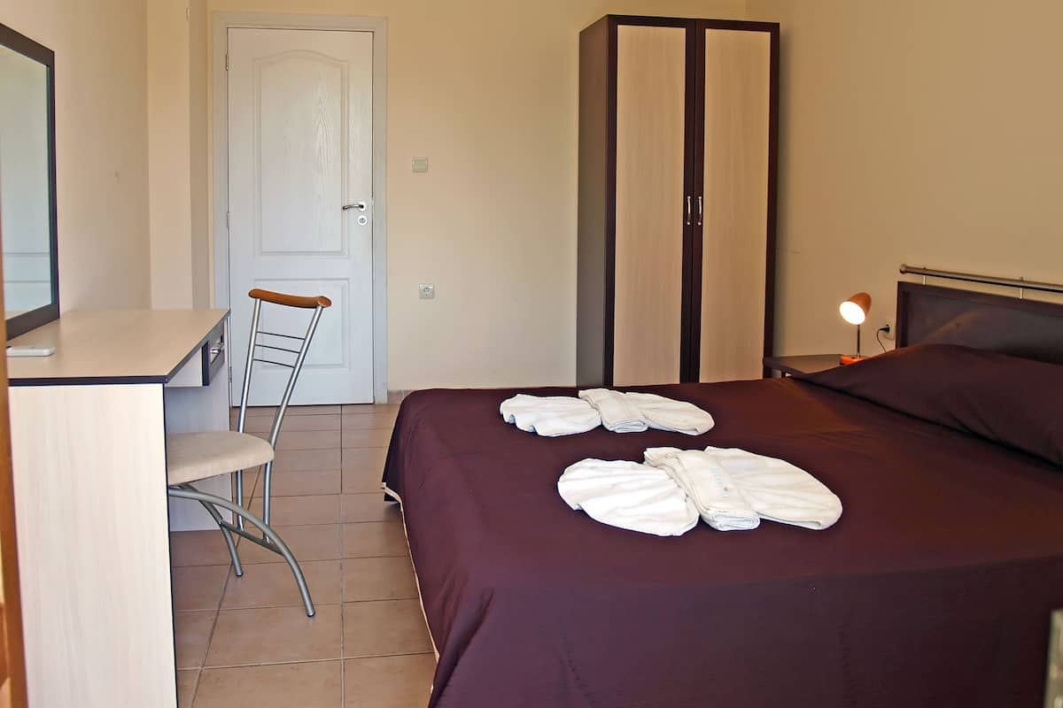 A hotel room with a bed, a desk, a chair, a wardrobe, and a bedside table with a lamp.