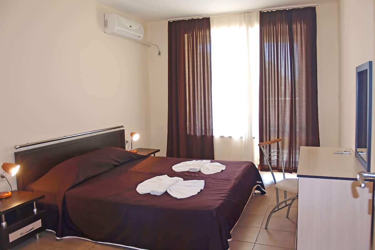 A hotel room with a bed, bedside tables, a desk, a chair, an air conditioning unit, and a window with curtains.