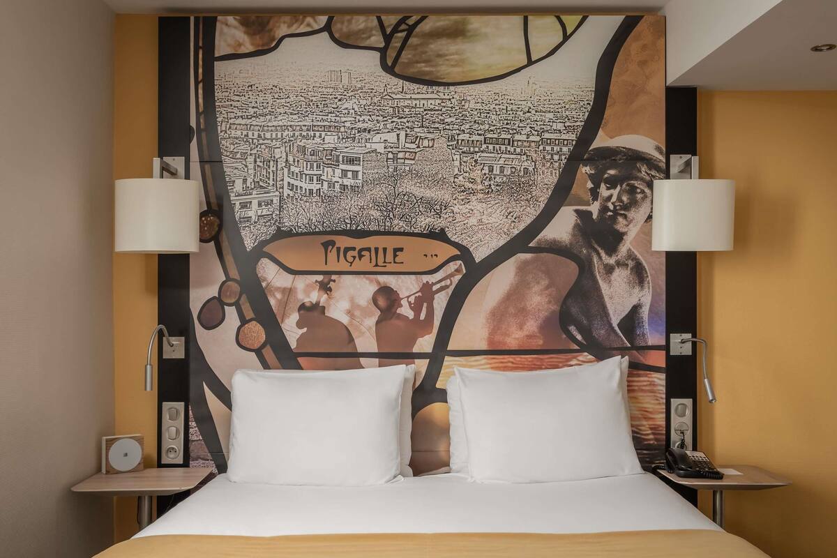 A hotel room with a bed, two bedside lamps, a nightstand with a phone, and a large wall mural.