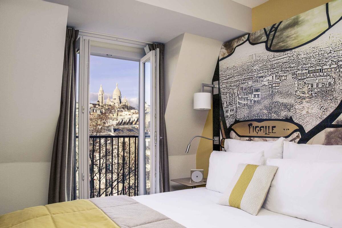 A hotel room with a bed, a window with curtains, a cityscape view, and a wall with a cityscape mural.