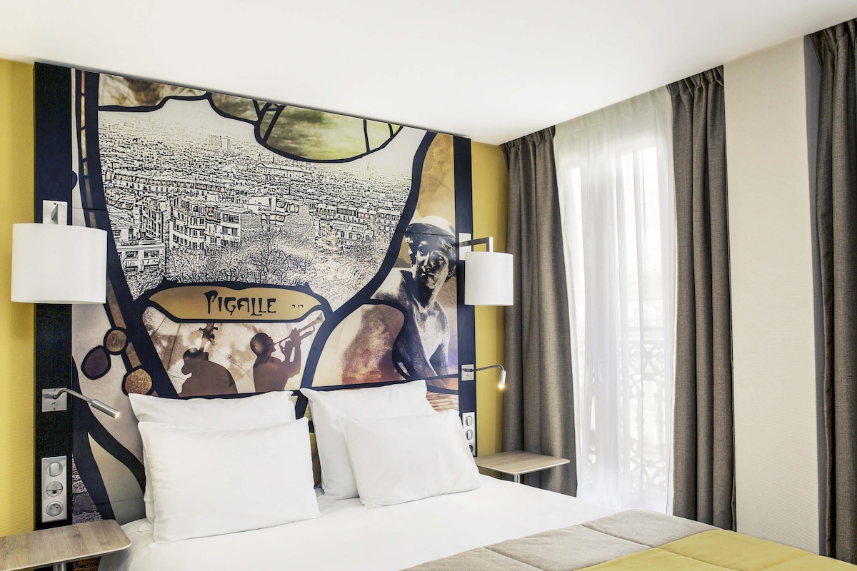 Une chambre d’hôtel avec un lit, deux tables de chevet, une fresque murale et une fenêtre avec des rideaux.
