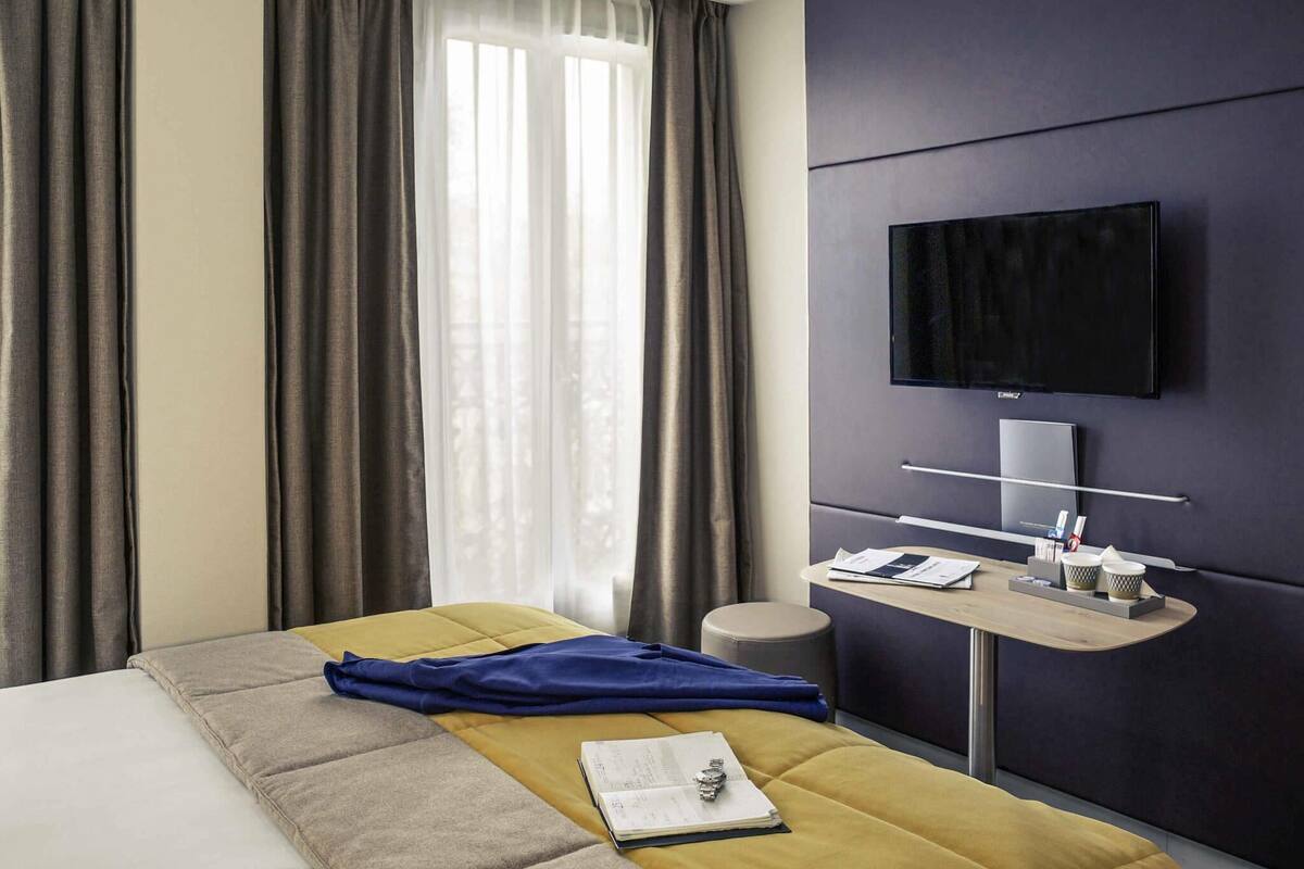 A hotel room with a bed, a desk with a flat-screen TV, a window with curtains, and a small round stool.