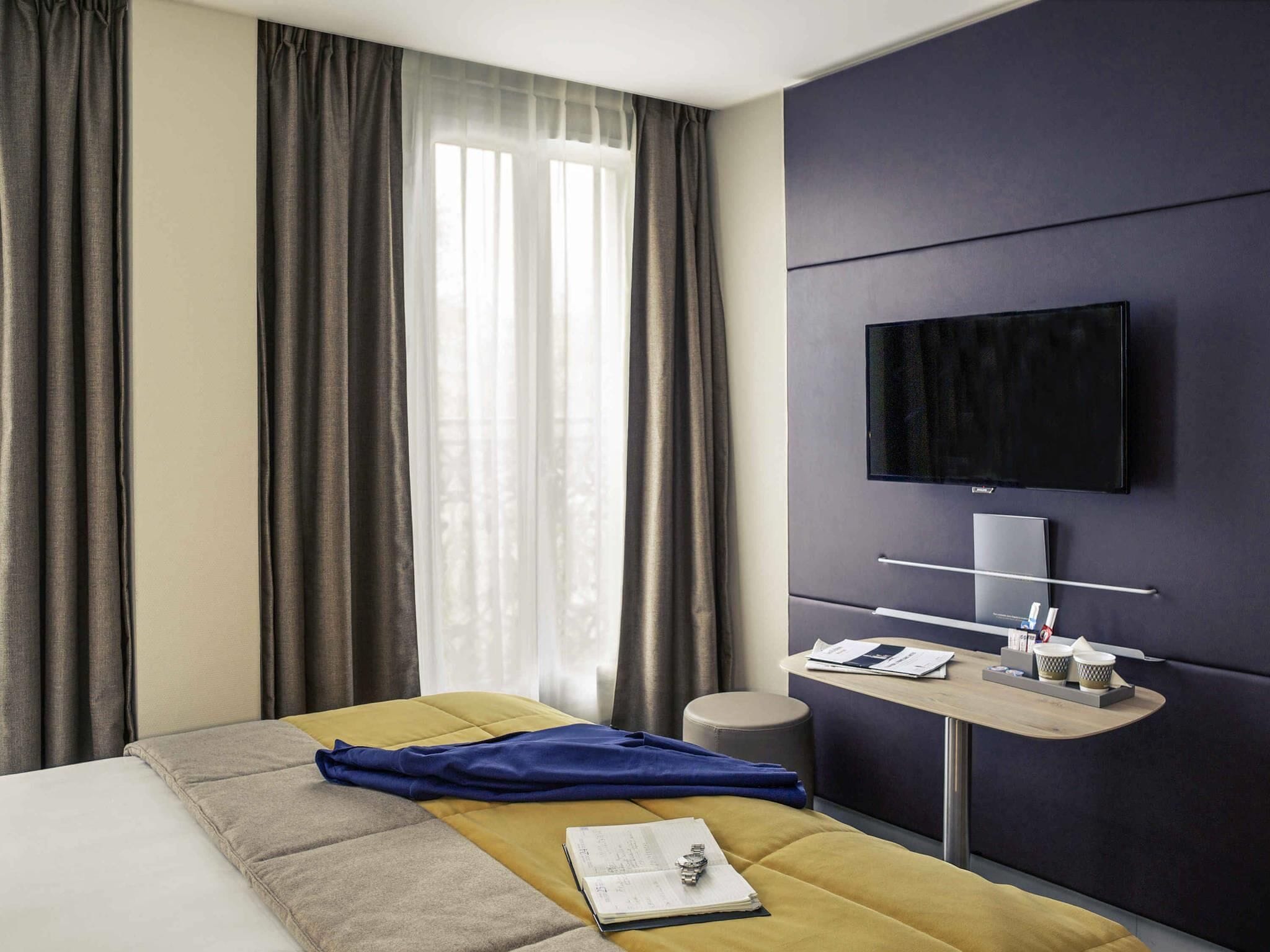 Une chambre d’hôtel comprenant un lit, un bureau avec un téléviseur à écran plat, une fenêtre avec des rideaux et un petit tabouret.