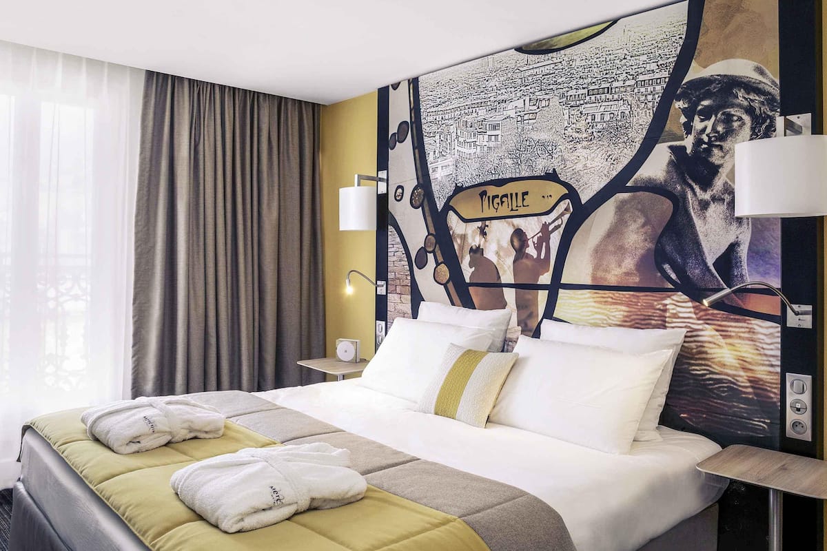 Une chambre d’hôtel avec un lit, des oreillers, une table de chevet, une lampe et une grande fresque murale.