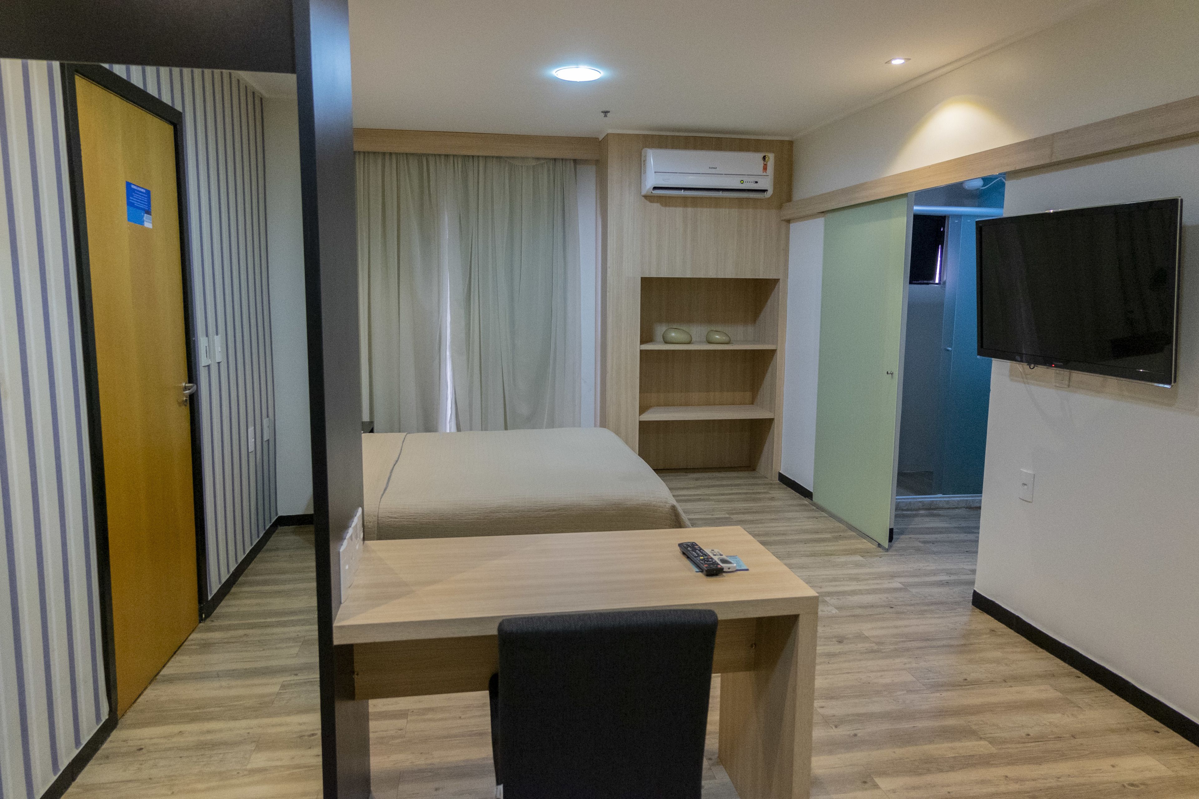 Habitación de hotel con cama, escritorio, silla, televisor y estante con artículos de aseo.