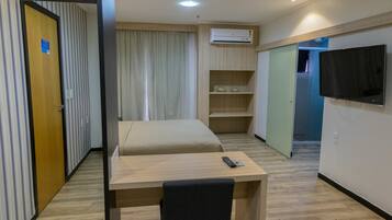 Habitación de hotel con cama, escritorio, silla, televisor y estante con artículos de aseo.