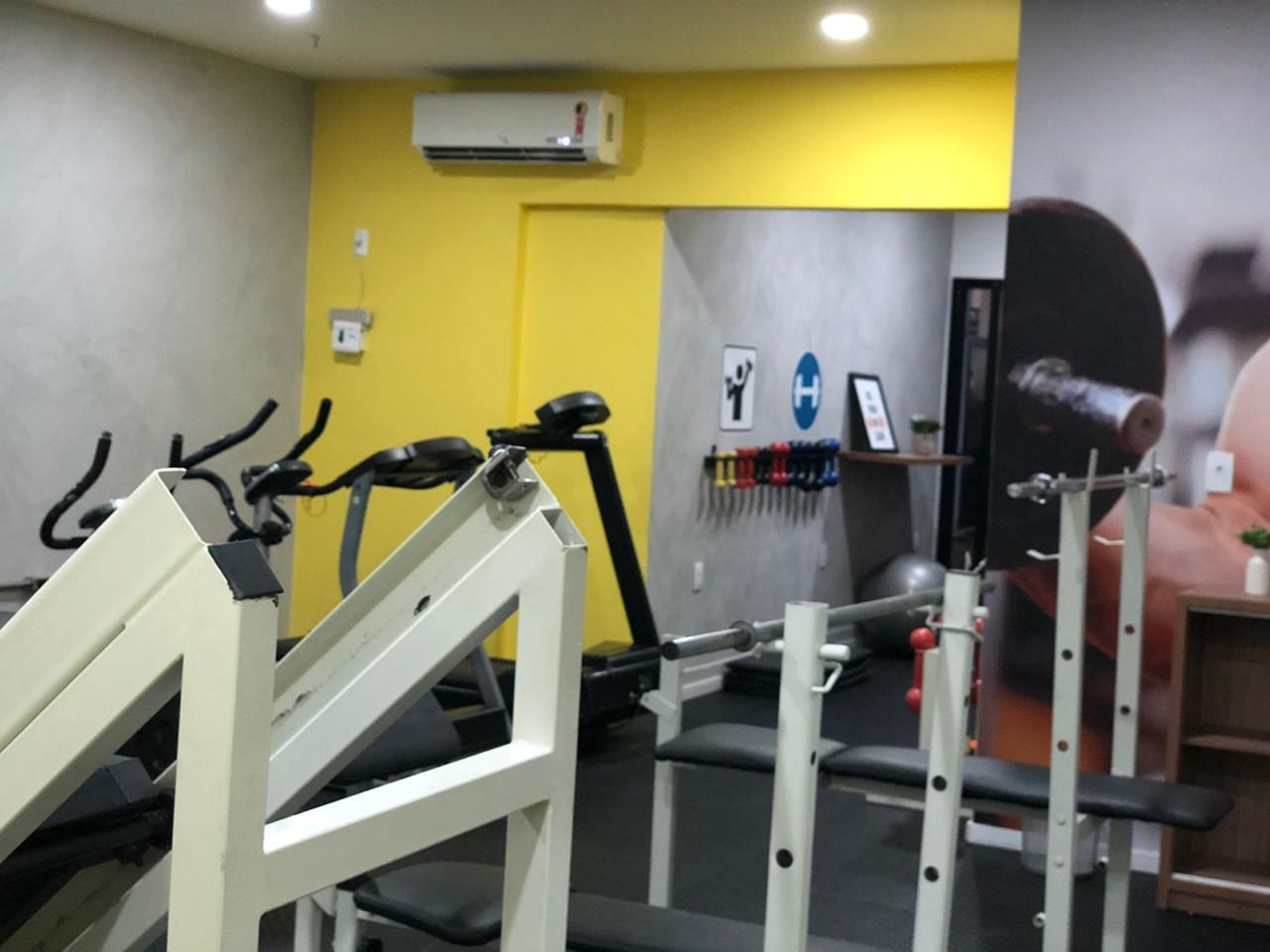 A gym with exercise equipment, a wall-mounted air conditioner, and a wall with a hand symbol and a blue 'H' sign.