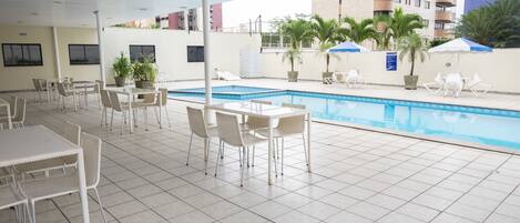 Zona de piscina interior con suelo de baldosas, sillones blancos y mesas.