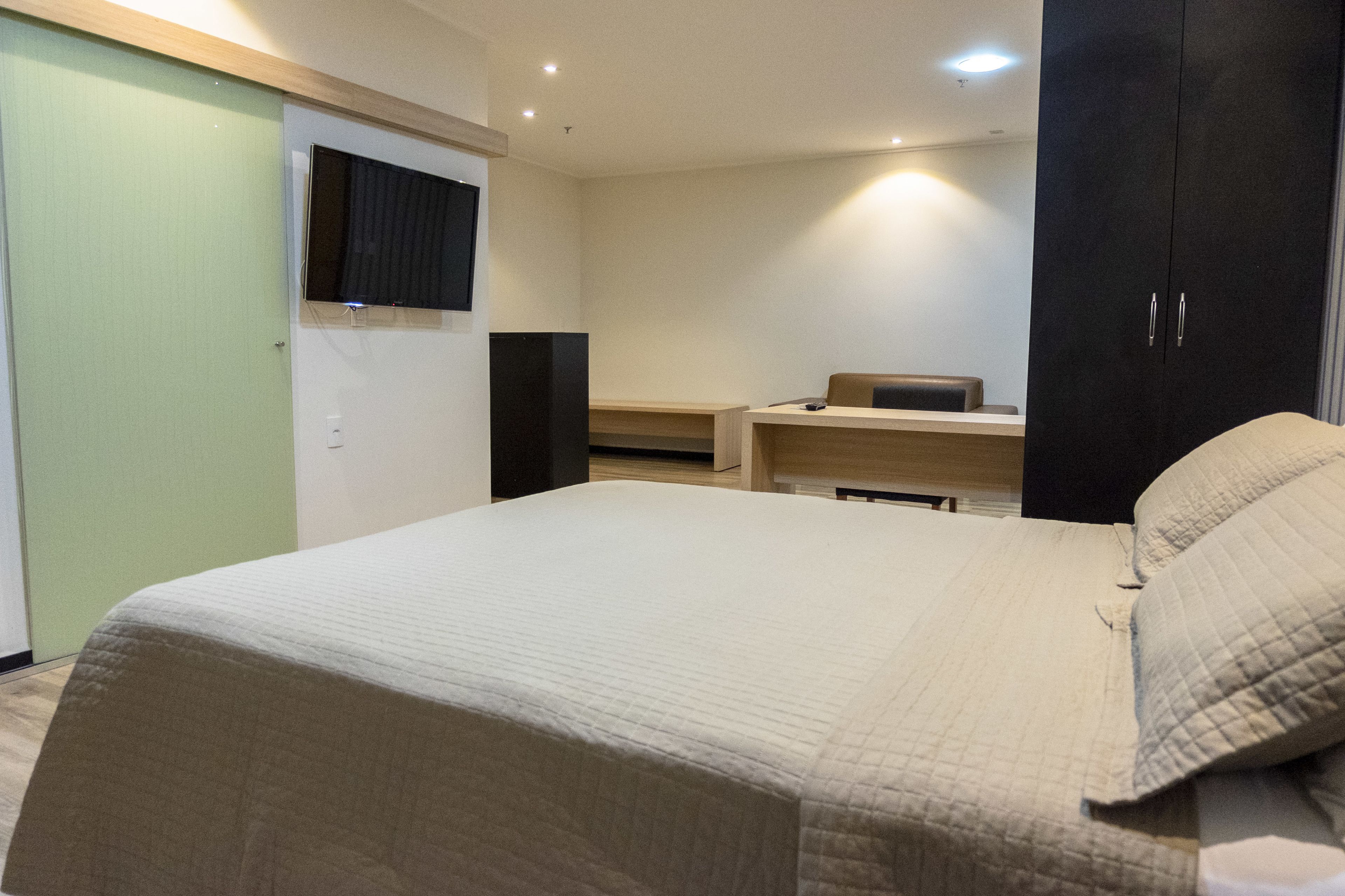 A bedroom with a bed, a TV mounted on the wall, a desk, and a wardrobe.