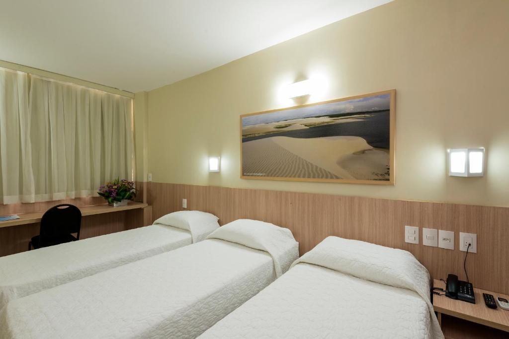 Une chambre d’hôtel avec deux lits simples, un bureau, une chaise, un tableau représentant un paysage désertique et une fenêtre avec des rideaux.