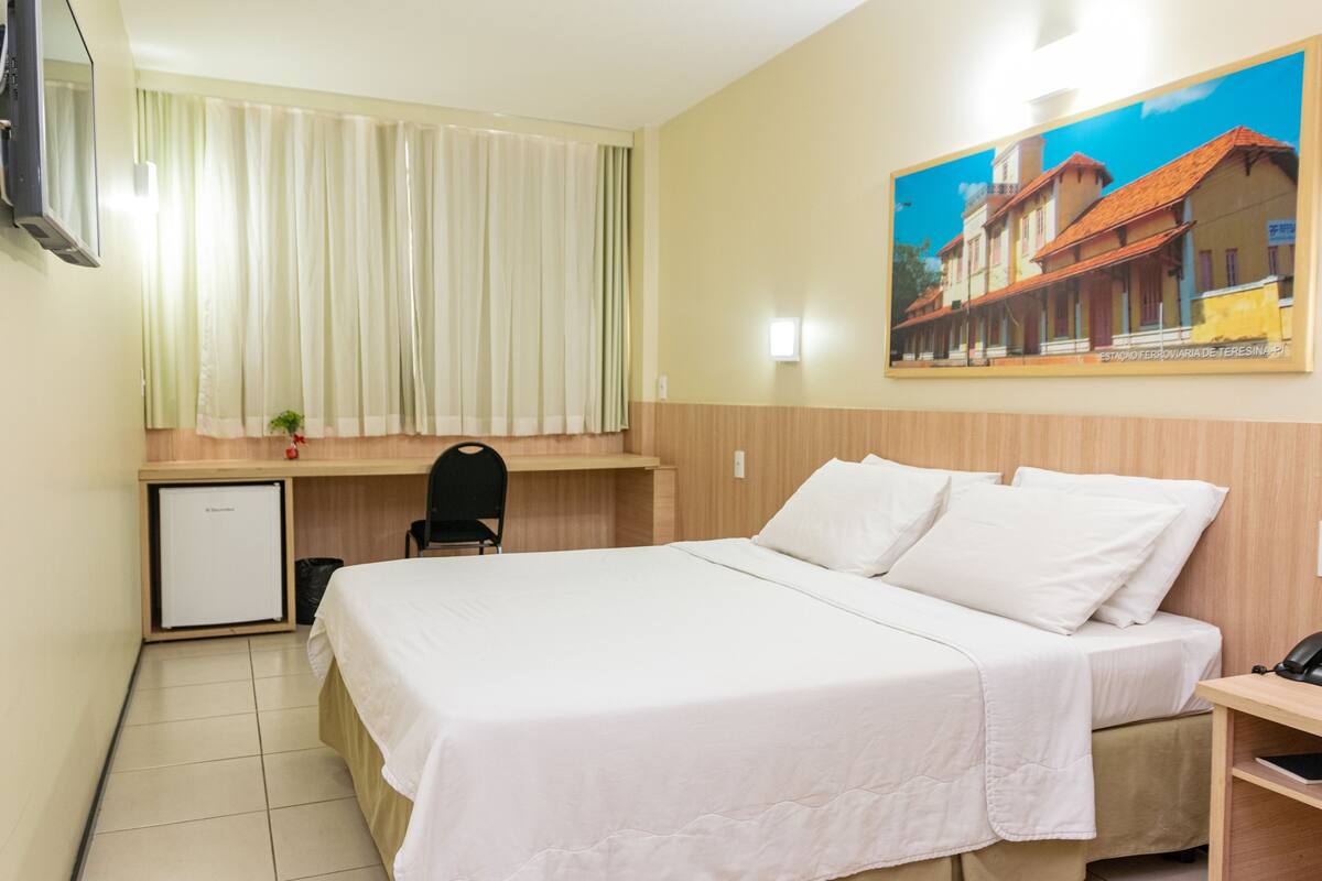 A hotel room with a bed, a desk, a chair, a small refrigerator, and a painting on the wall.