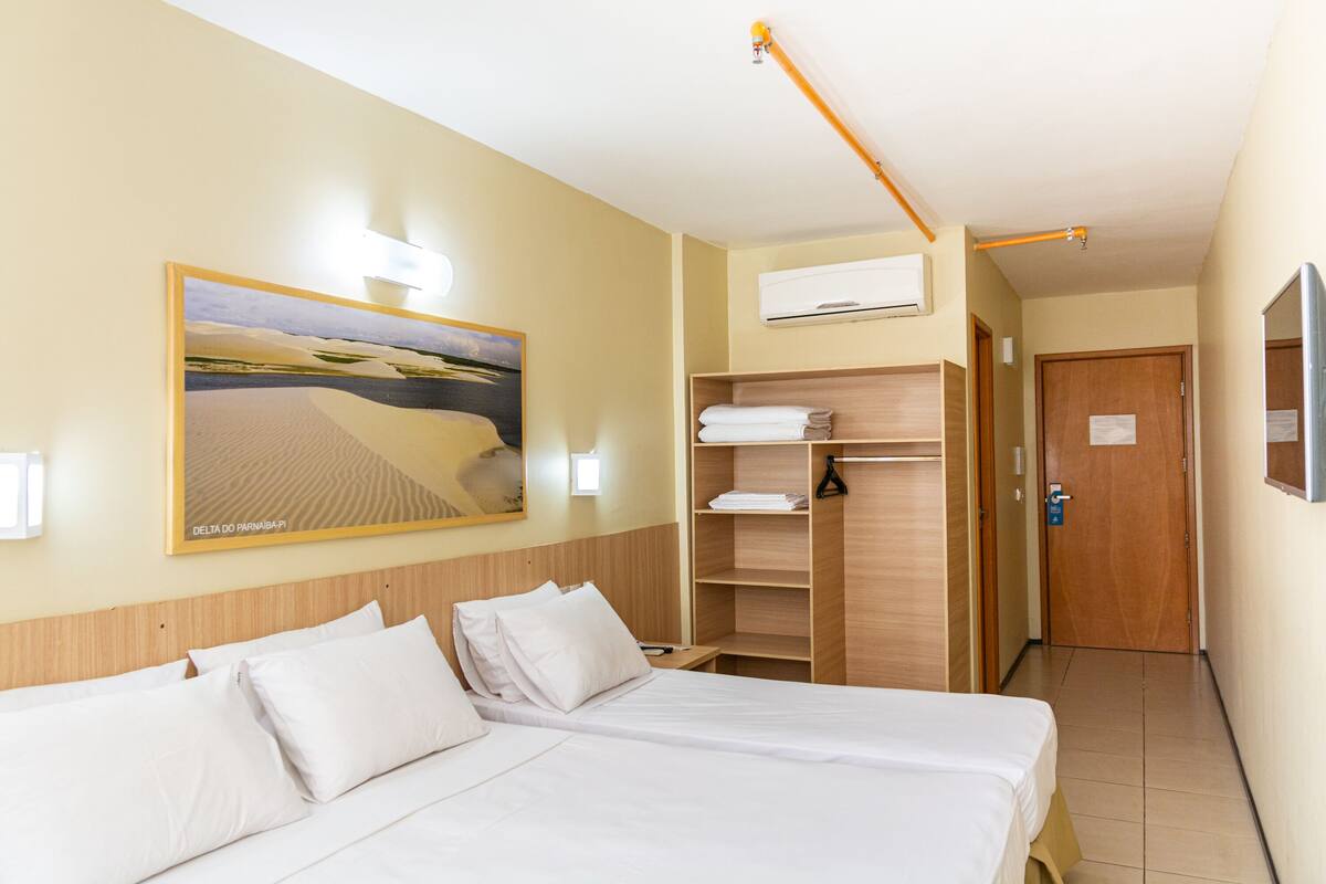 A hotel room with a bed, a wooden headboard, a painting of a desert landscape, a wardrobe, and an air conditioning unit.