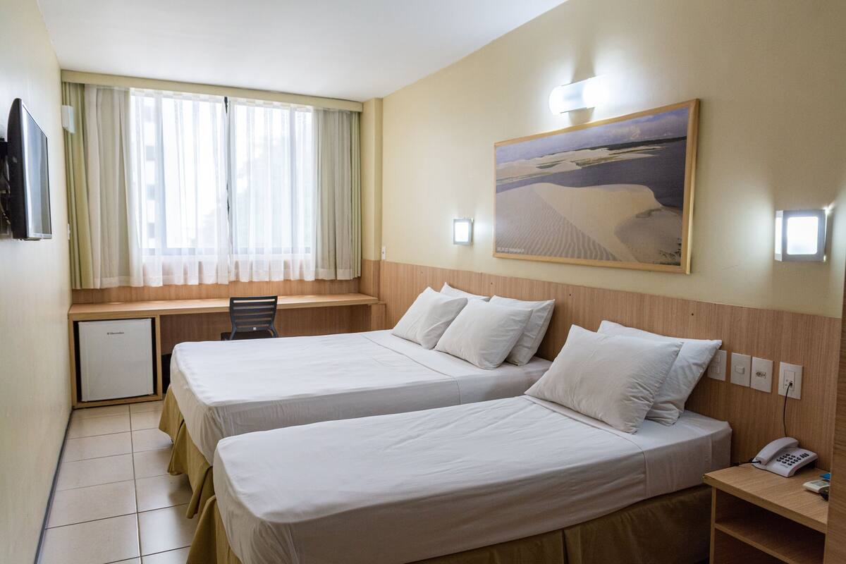 A hotel room with two beds, a desk, a chair, a small fridge, and a painting on the wall.