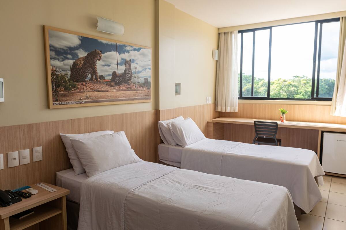 A hotel room with two beds, a desk, a chair, a small refrigerator, and a large window with a view of trees.