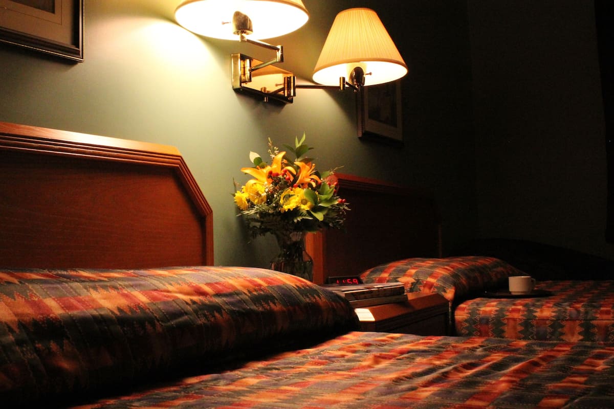 Habitación de hotel con dos camas, una mesita de noche con lámpara, un jarrón con flores y un reloj.