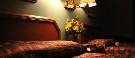 Habitación de hotel con dos camas, una mesita de noche con lámpara, un jarrón con flores y un reloj.