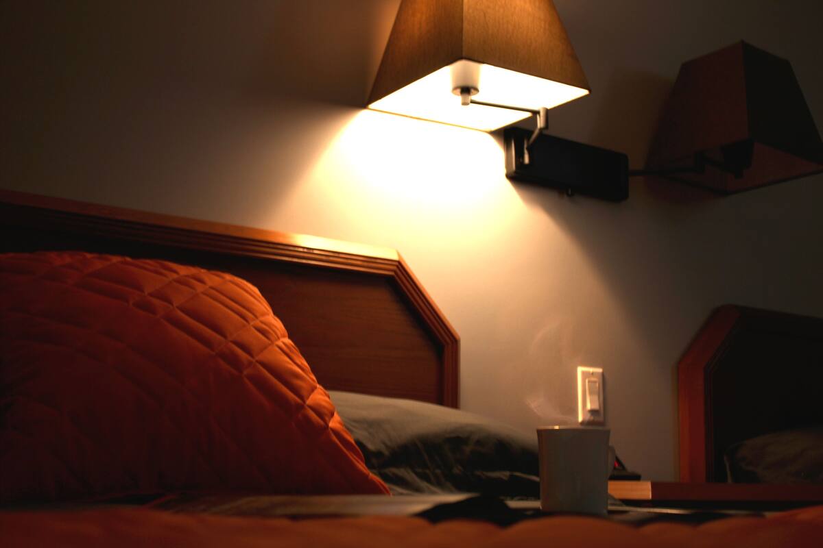 Habitación de hotel con poca luz, una cama, una lámpara de noche y un mesita de noche con una taza.