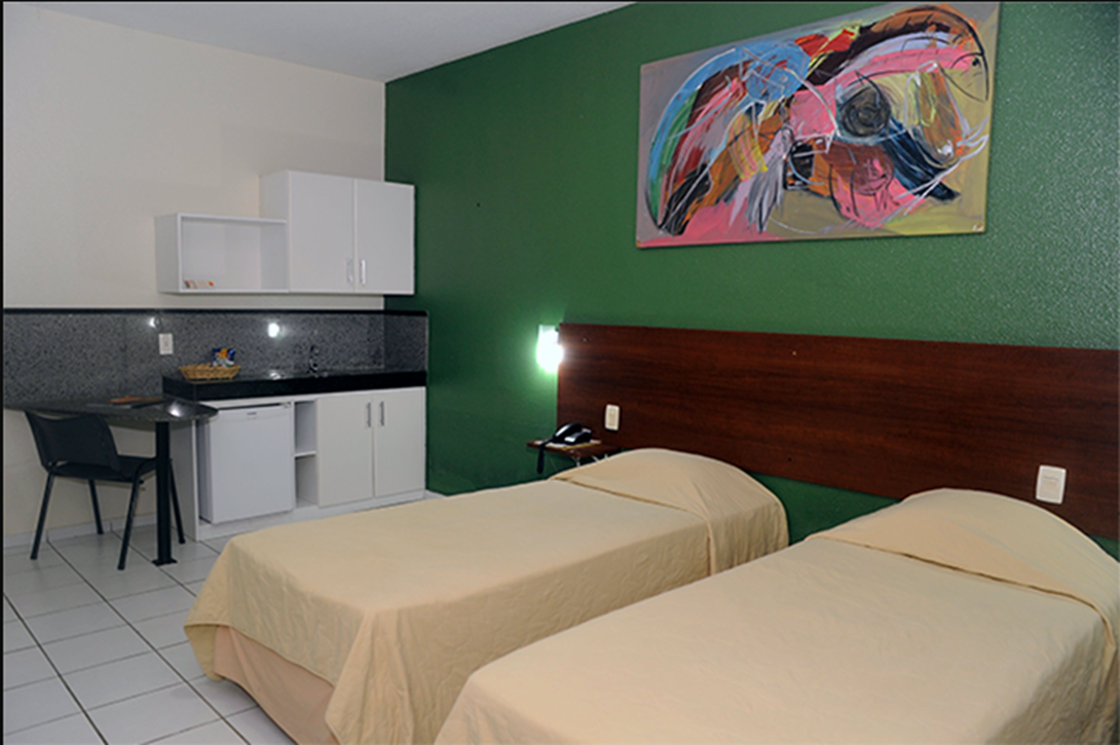 A hotel room with two beds, a desk, a chair, a small kitchenette, and a painting on the wall.