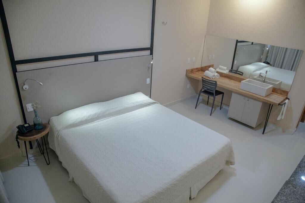 A hotel room with a bed, a desk with a chair, a mirror, and a small table with a vase.