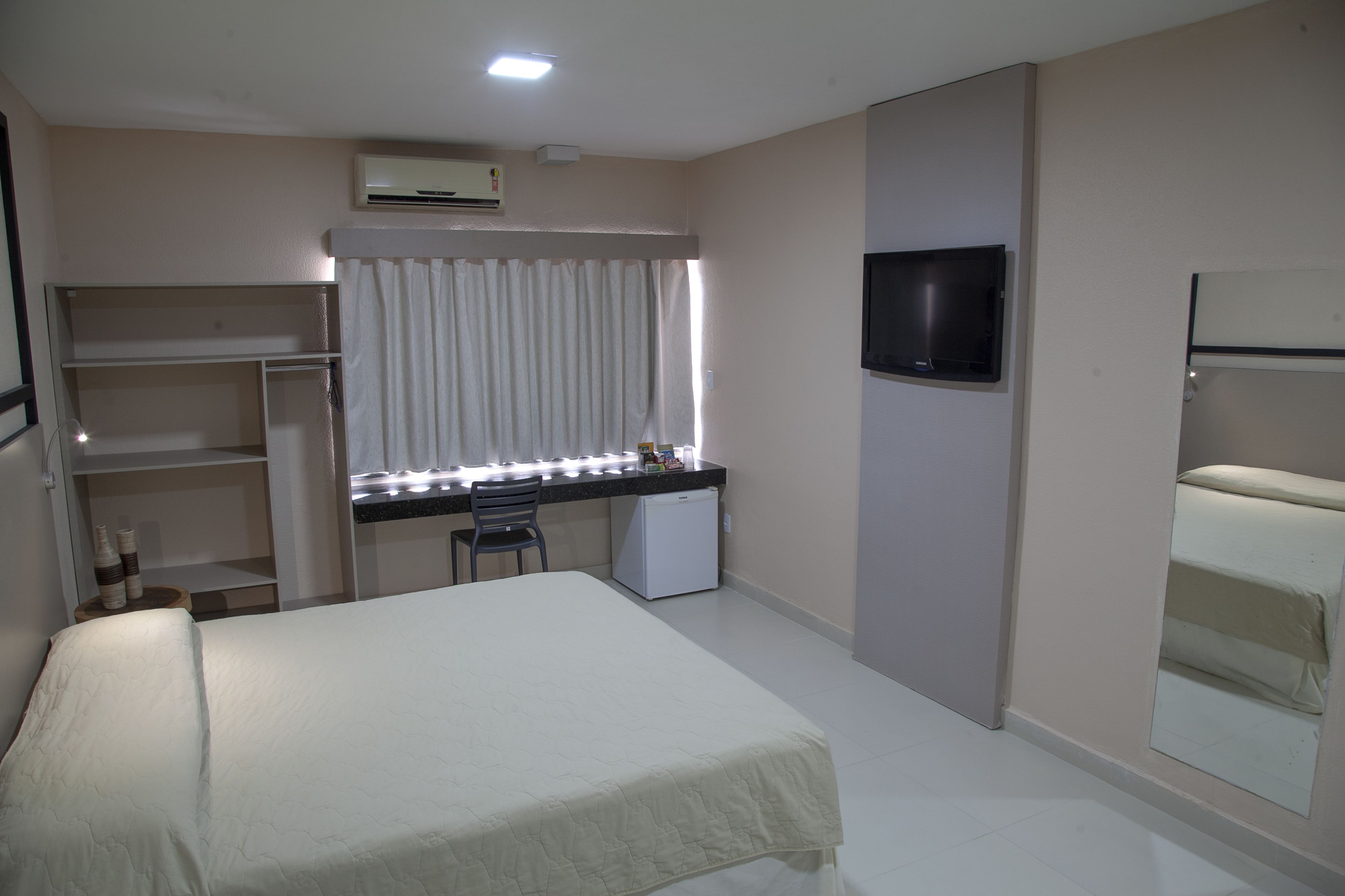 A hotel room with a bed, a desk with a chair, a small refrigerator, a TV mounted on the wall, and a window with curtains.