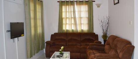 A living room with a brown sofa, a white coffee table, and a TV on the wall.
