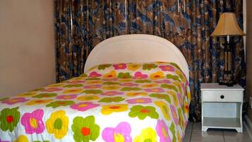Ein Bett mit floralem Überwurf, ein Nachttisch mit Lampe und Vorhänge mit Blattmuster.