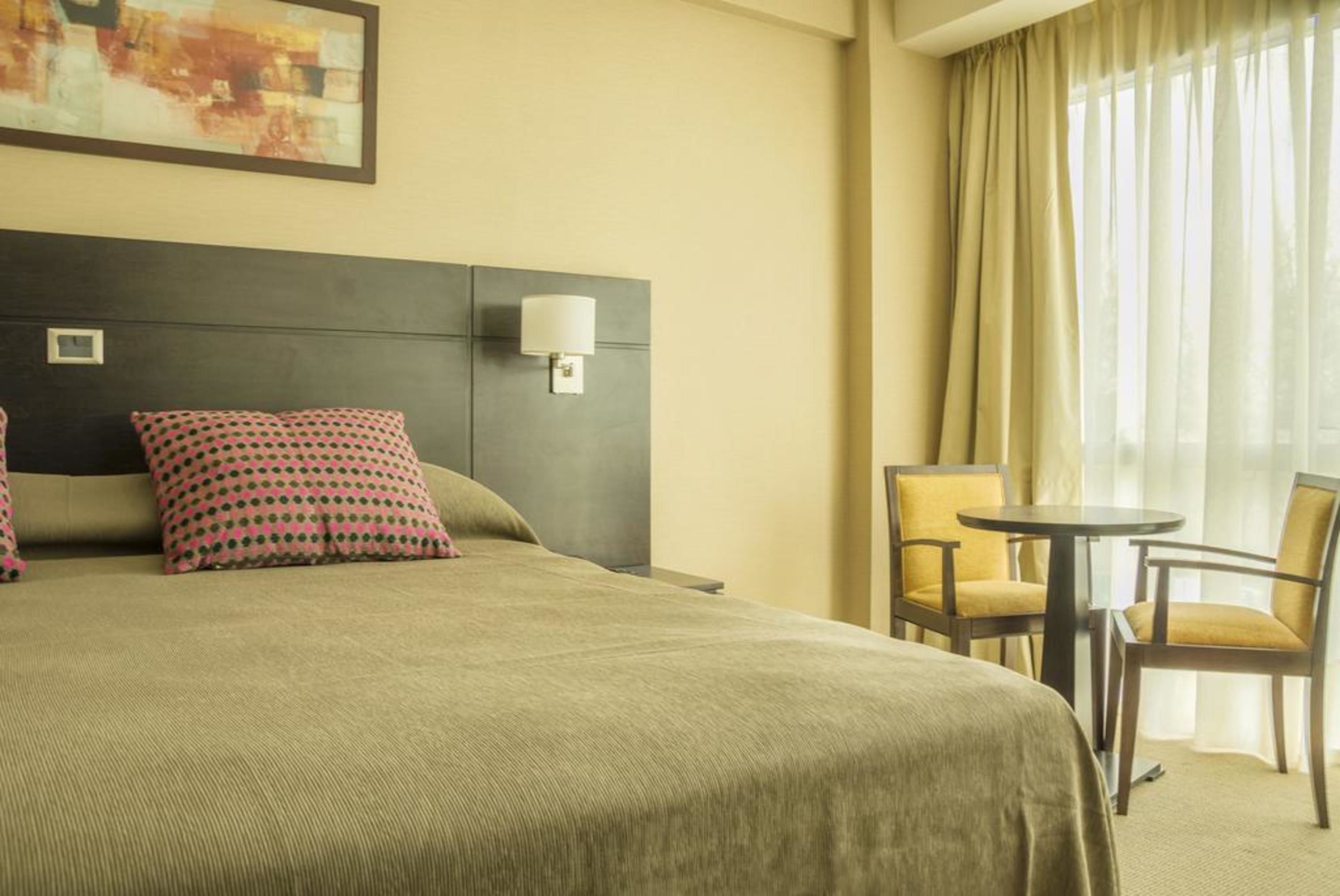 A hotel room with a bed, two pillows, a nightstand, a lamp, a chair, and a table.
