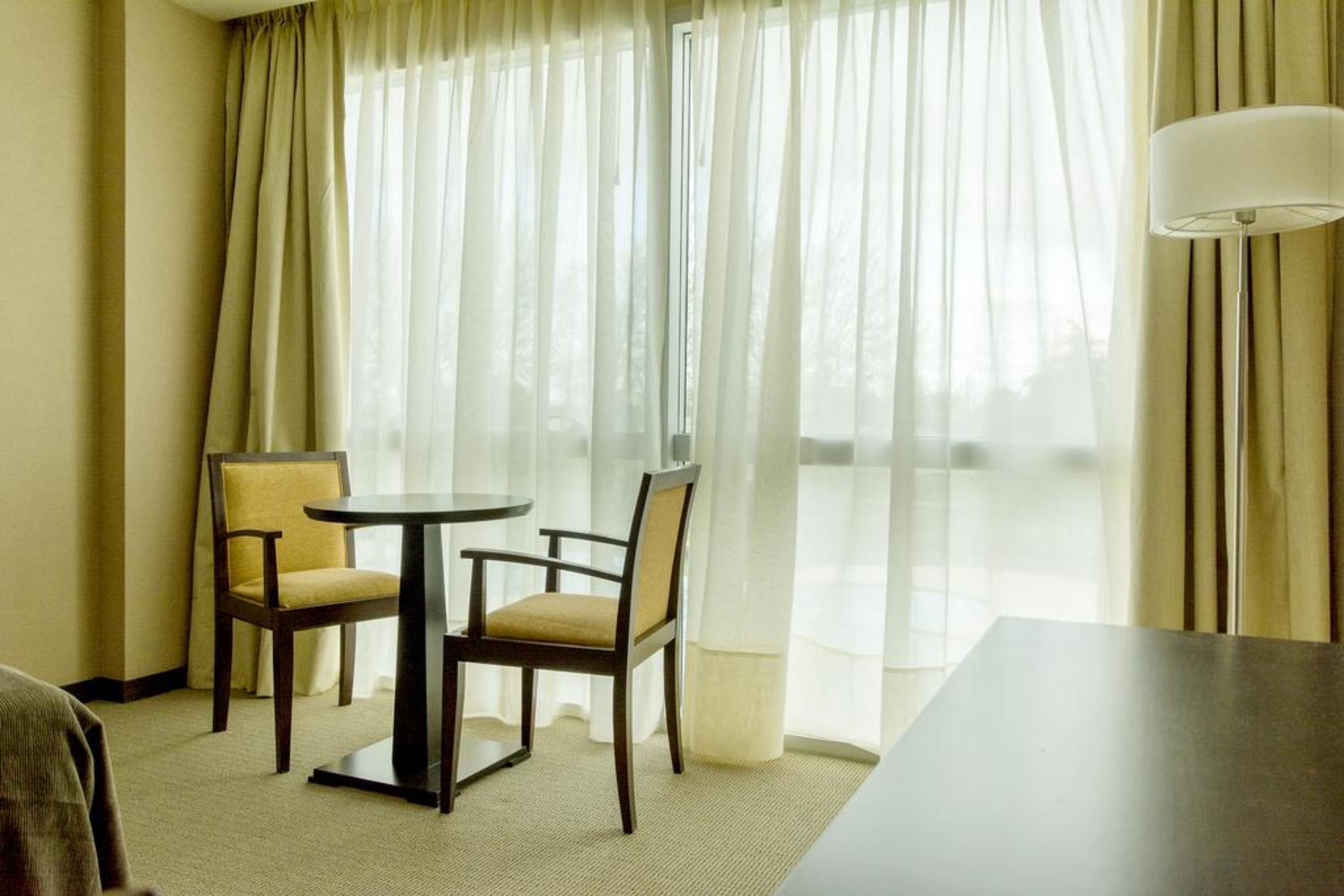 Ein Hotelzimmer mit Essbereich, ausgestattet mit zwei Stühlen und einem Tisch, einem großen Fenster mit durchsichtigen Vorhängen und einer Lampe auf der rechten Seite.