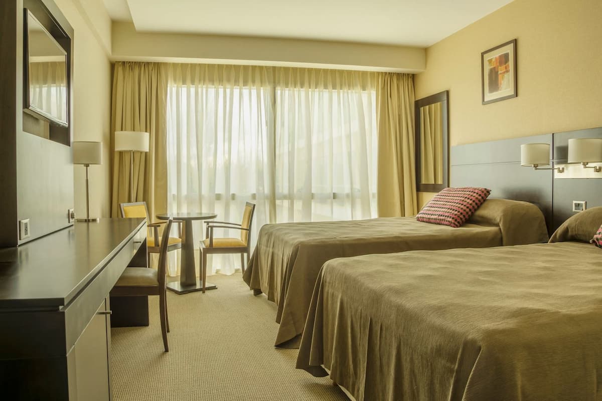 Une chambre d’hôtel avec deux lits, un bureau, une chaise et une grande fenêtre avec des rideaux.