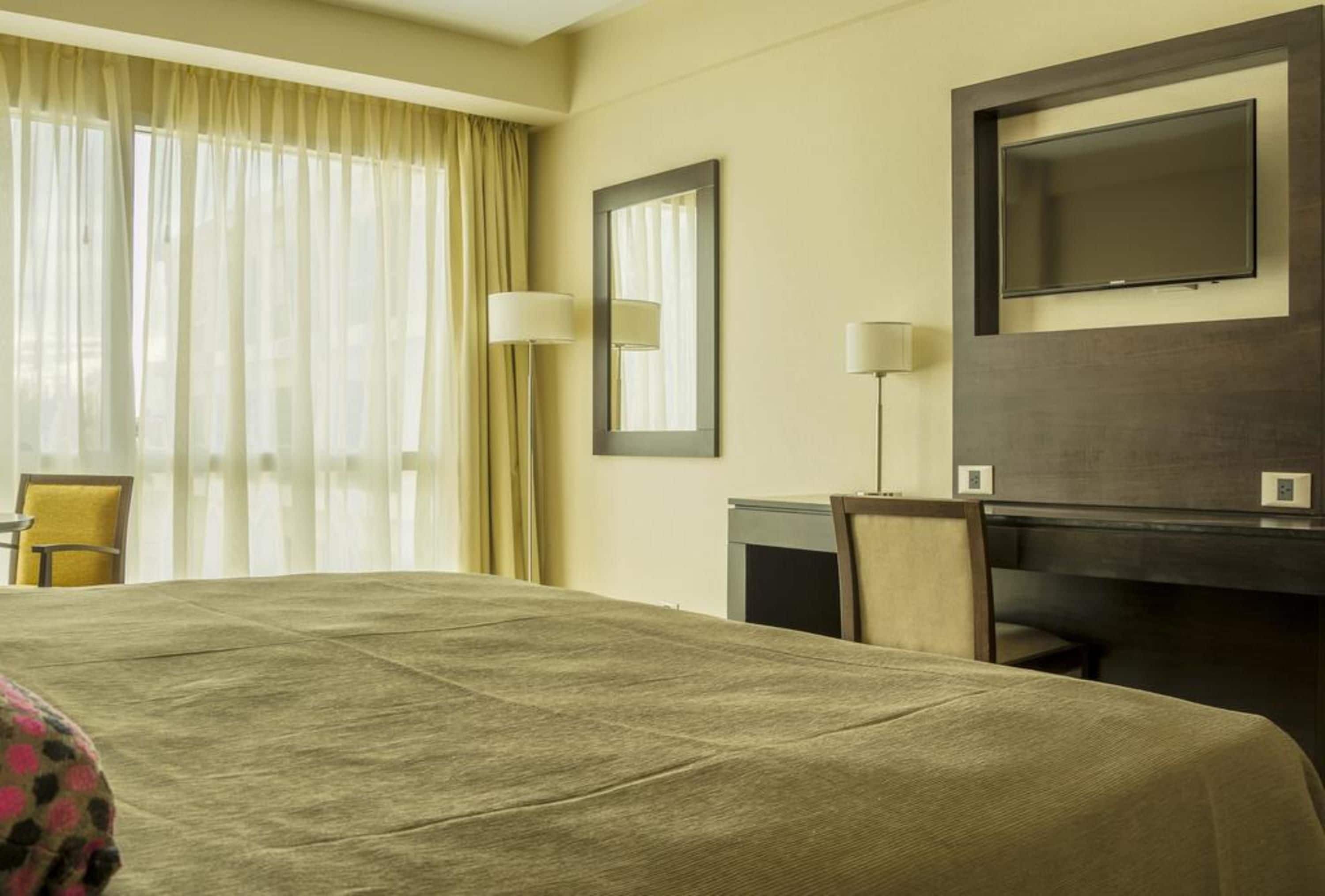 A hotel room with a large bed, a desk, a chair, a TV, and a window with curtains.