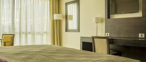 A hotel room with a large bed, a desk, a chair, a TV, and a window with curtains.