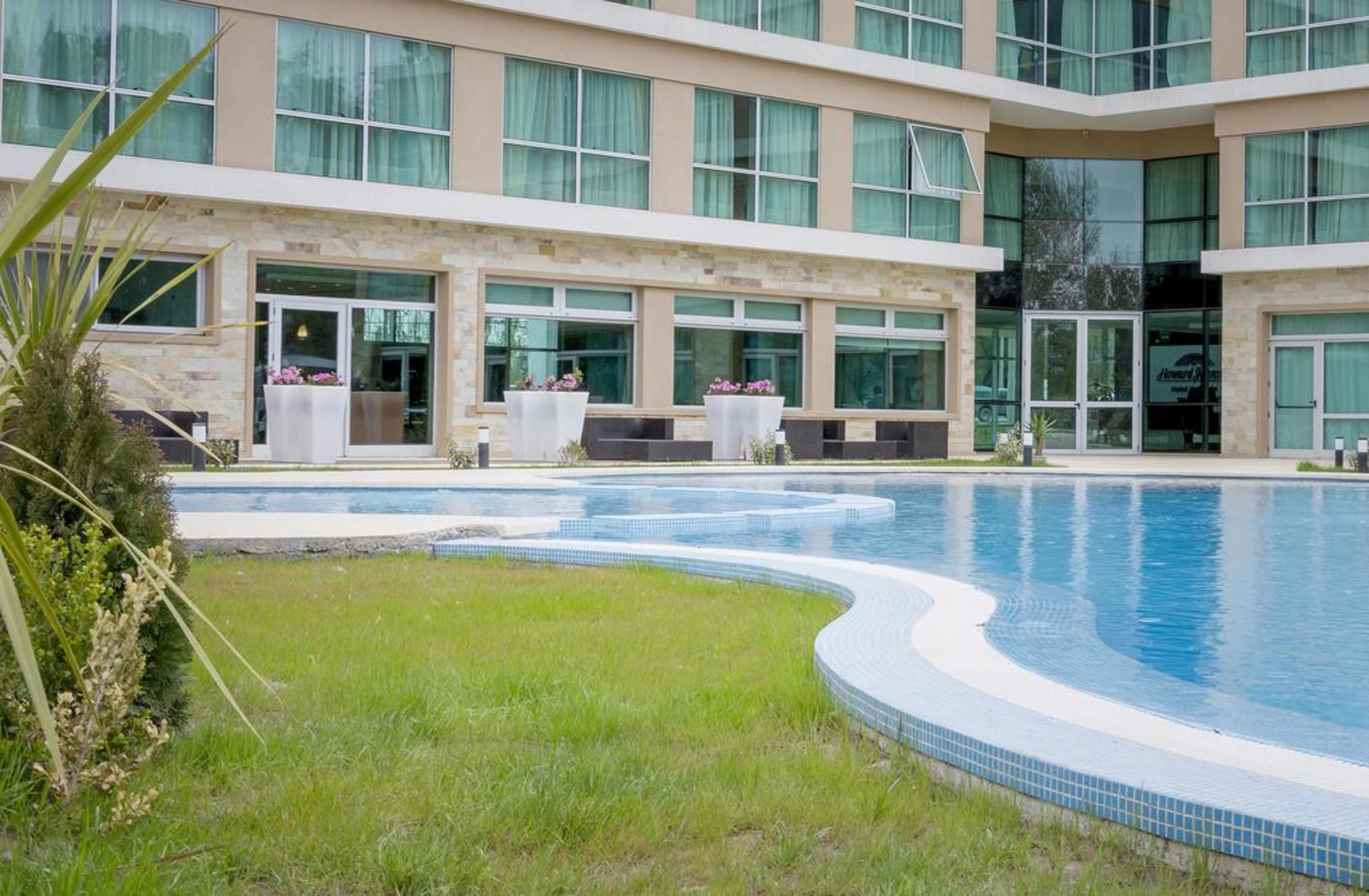 A modern hotel with a large swimming pool, outdoor seating, and a well-maintained garden area.