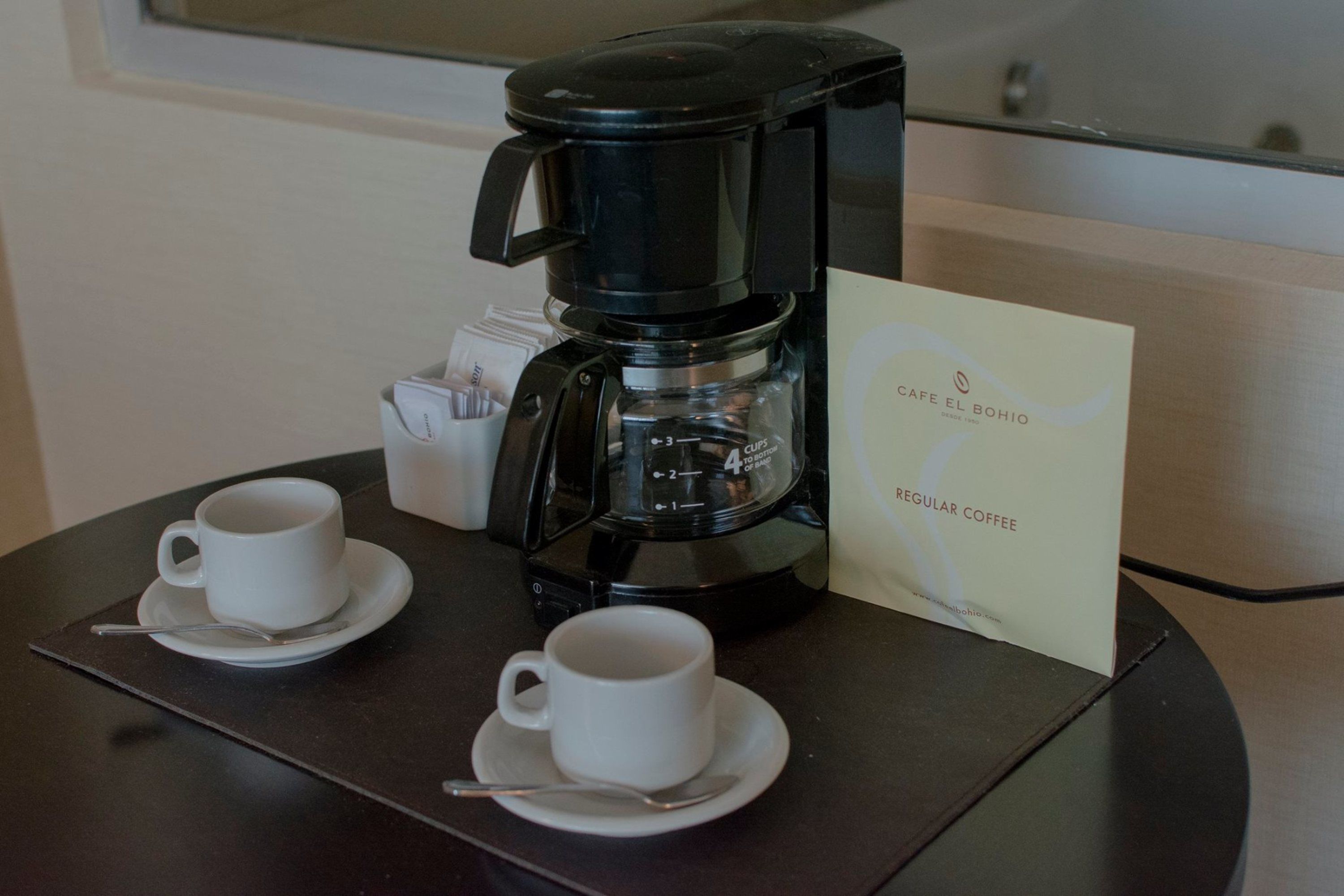 Een koffiezetapparaat met twee kopjes op een tafel, een bord met de tekst 'Café El Bohio' en een lepel.