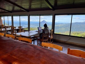 A dining area with a table and chairs, a person working on a laptop, and large windows offering a view of a mountainous landscape.