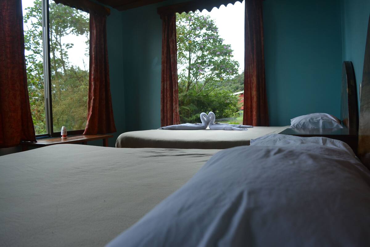 A room with two beds, a window with curtains, and a view of trees and a house outside.
