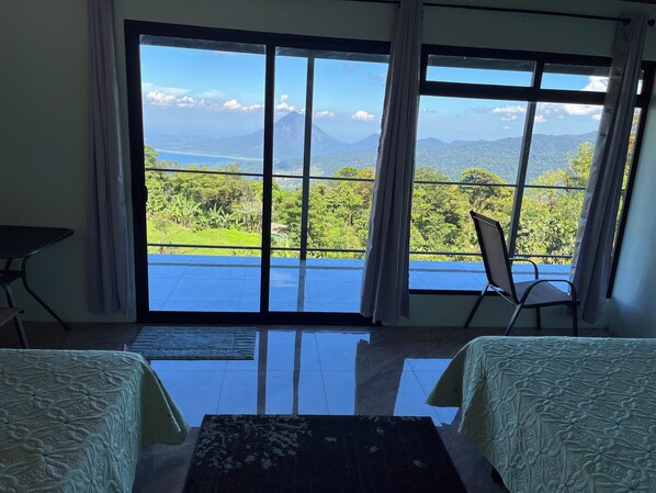 A room with a large sliding glass door leading to a balcony with a view of a mountain and greenery. There are two chairs and a small table in the room.
