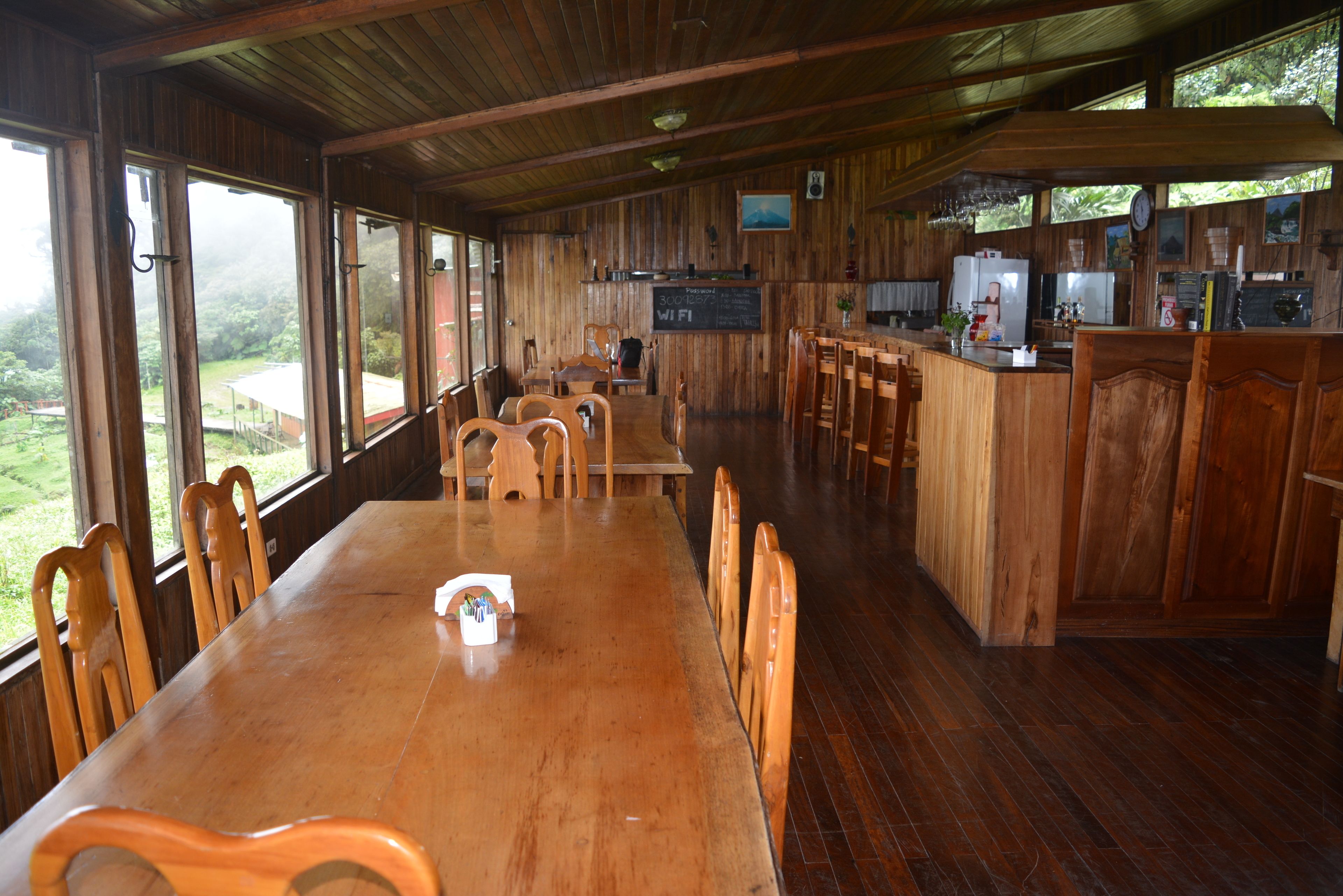 Un comedor de madera con una mesa larga y sillas, un área de cocina con barra, y ventanas amplias que ofrecen vistas al exterior.