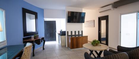 A modern hotel room with a dining area, a small kitchen, and a seating area with wicker chairs.