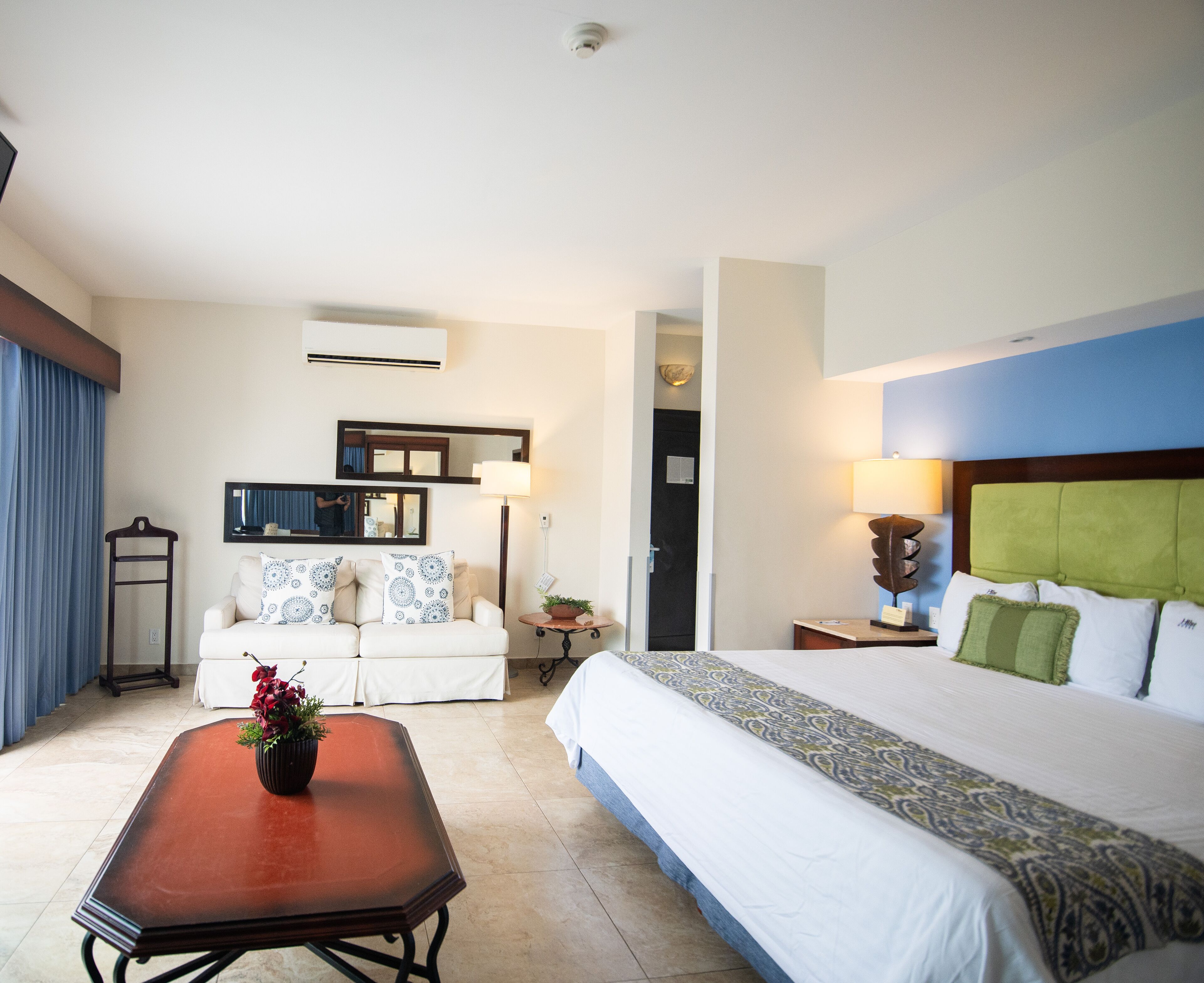 A hotel room with a bed, a sofa, a table with a vase, a mirror, and a lamp.