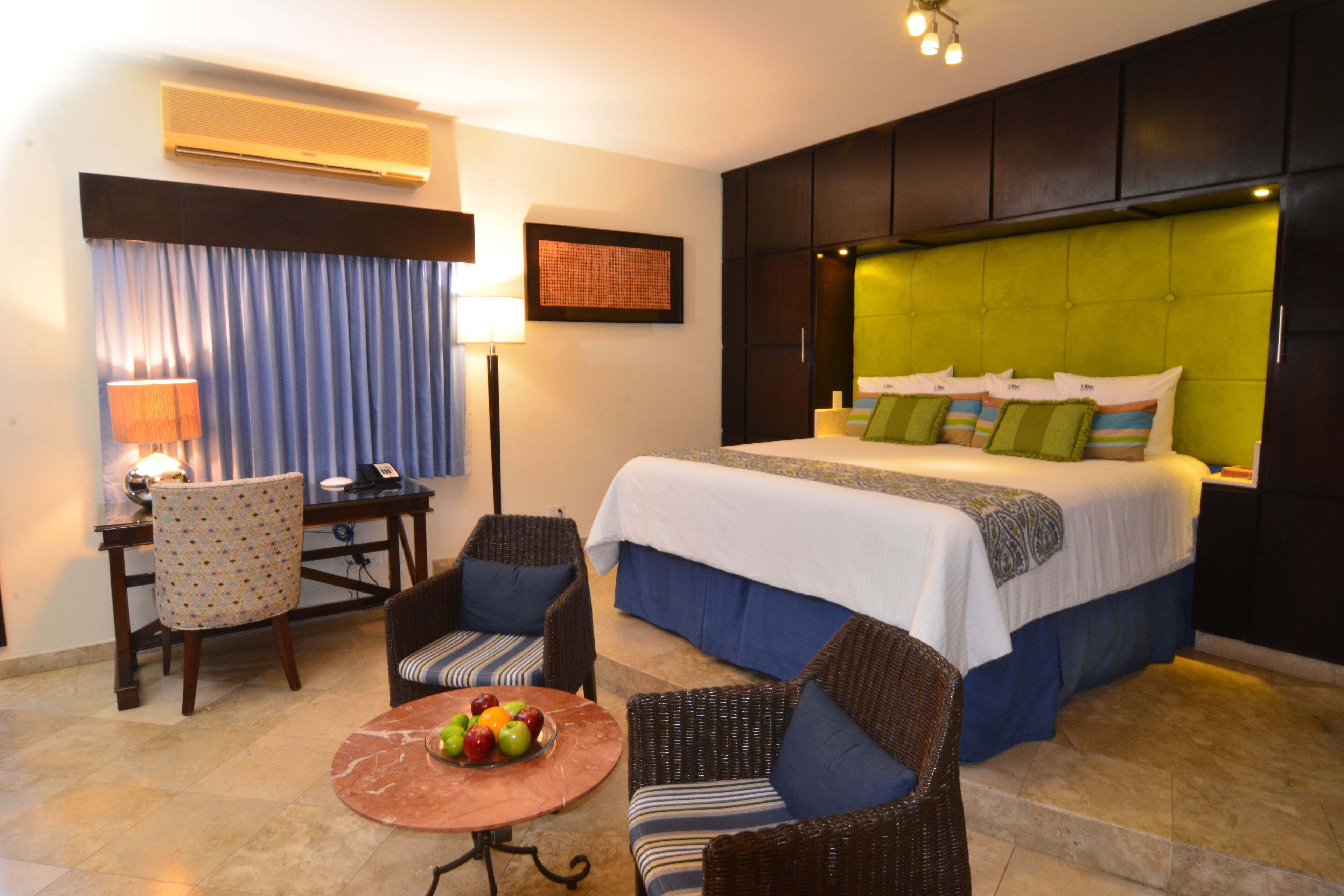 Habitación de hotel con una cama, dos sillas, una mesa con fruta, un escritorio y una lámpara.