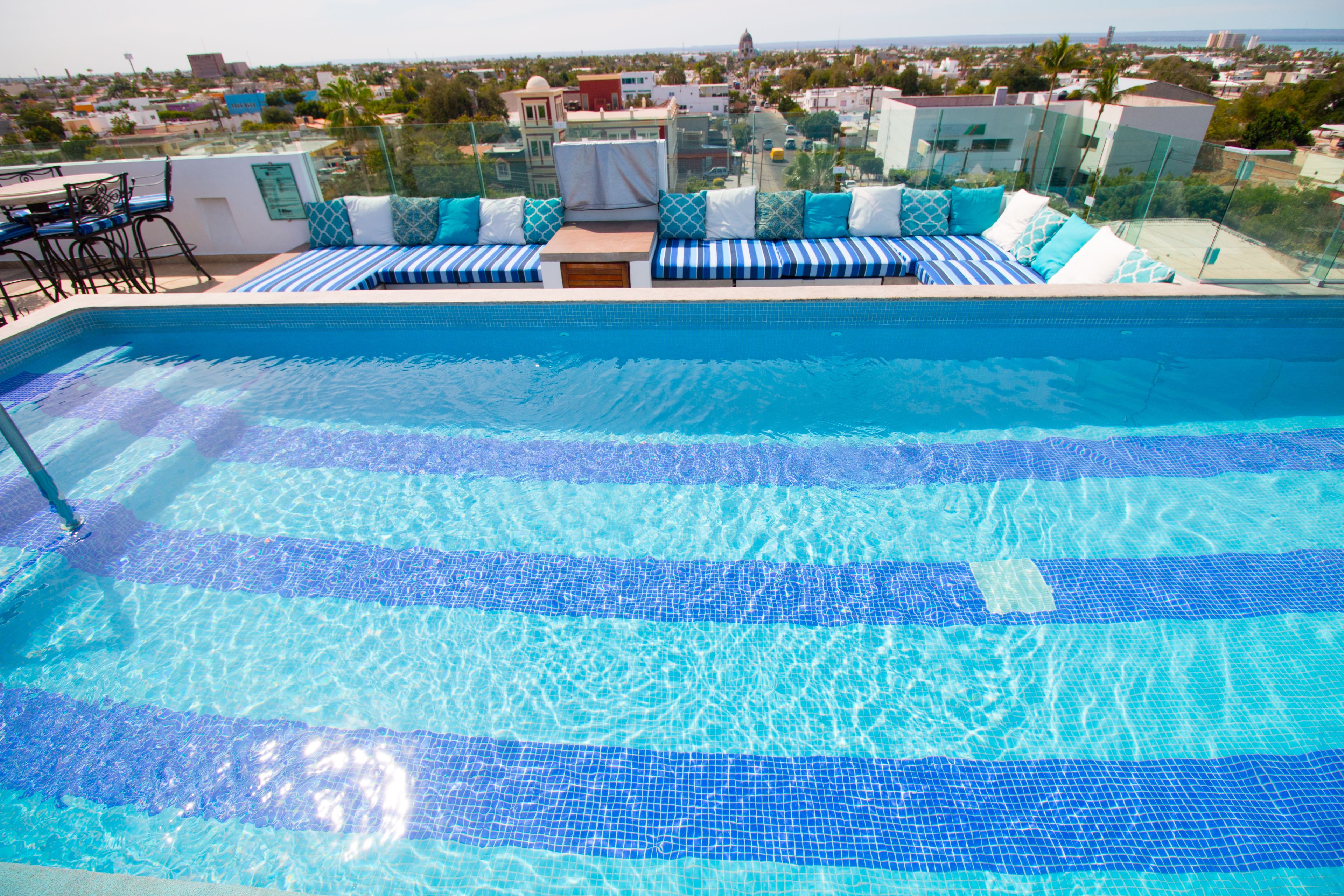 Une piscine d’un bleu éclatant, bordée d’une terrasse carrelée, un espace détente avec des coussins et, en arrière-plan, une vue sur le paysage urbain.