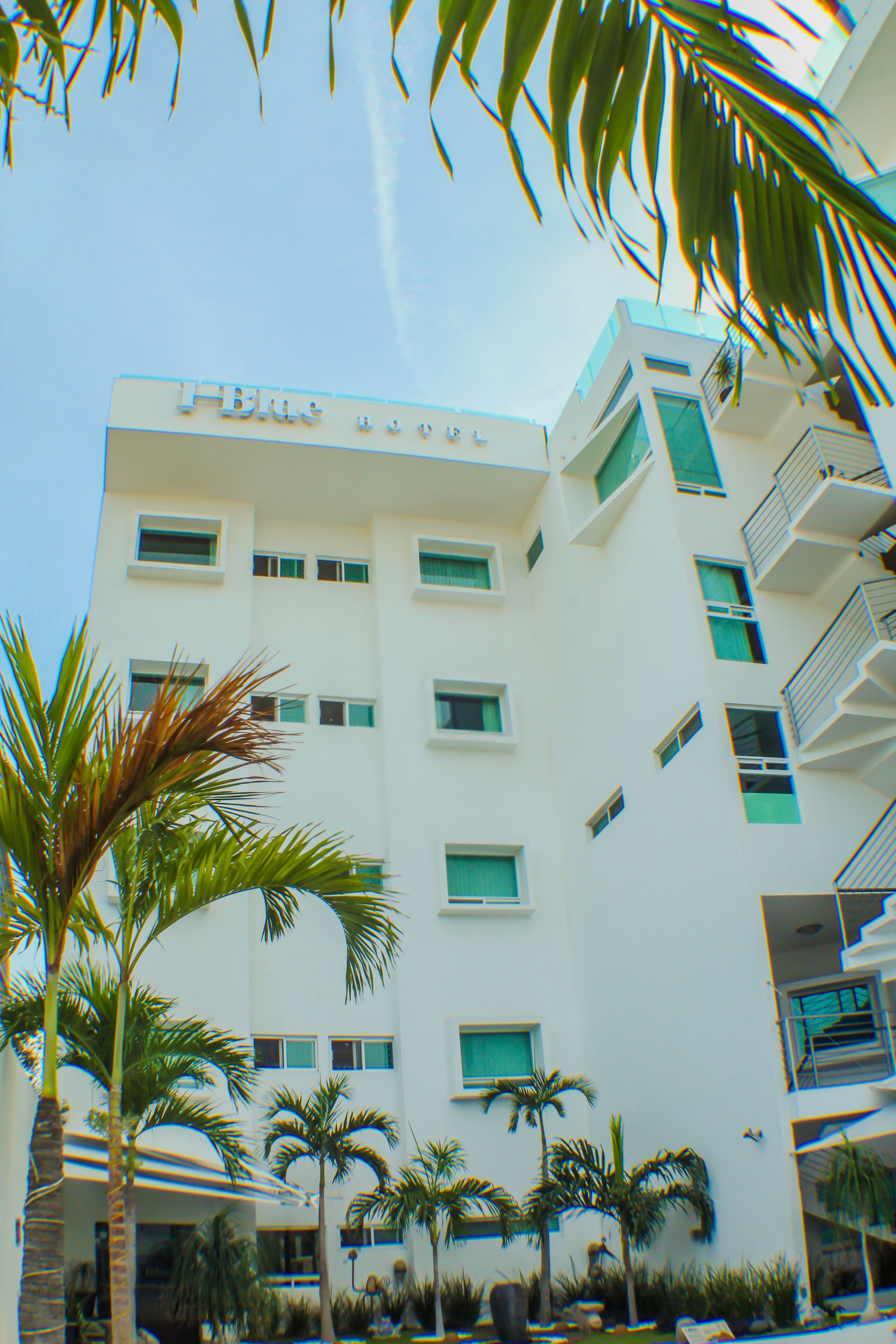 Ein mehrstöckiges Hotel mit weißer Fassade, mehreren Balkonen und Palmen im Vordergrund.