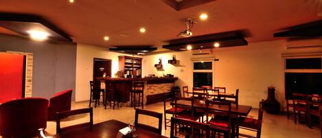 Uma ampla área de refeições com mesa e cadeiras de madeira, um espaço bar com banquetas e uma cozinha ao fundo.