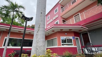 Um hotel com vários andares, fachada em vermelho e branco, varandas e área de entrada pavimentada com plantas.