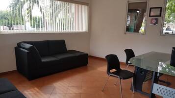 A room with a black sofa, a glass table with chairs, and a window with blinds.