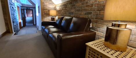 A cozy living area with a leather sofa, a wooden side table with a lamp, and a brick wall. A hallway leads to another room.