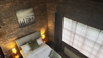 A bedroom with a brick wall, a bed with pillows, a nightstand with a lamp, and a window with blinds.
