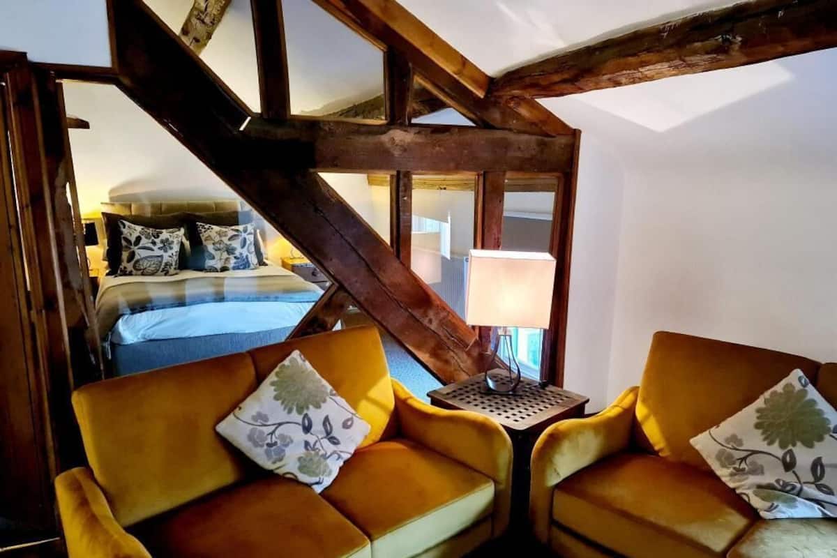 A room with a bed, two yellow sofas with floral cushions, a small table with a lamp, and exposed wooden beams.