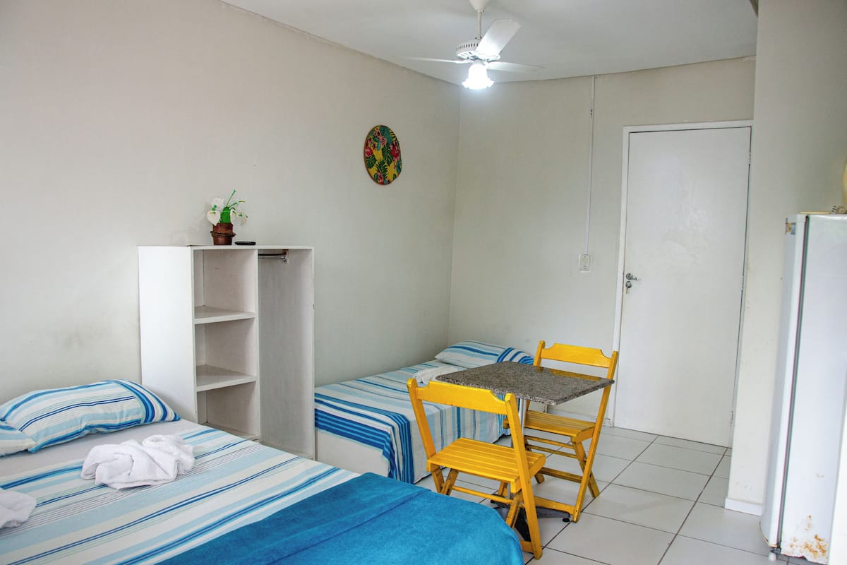 Quarto pequeno e limpo com duas camas, uma mesa, cadeiras, ventoinha no teto e uma porta.