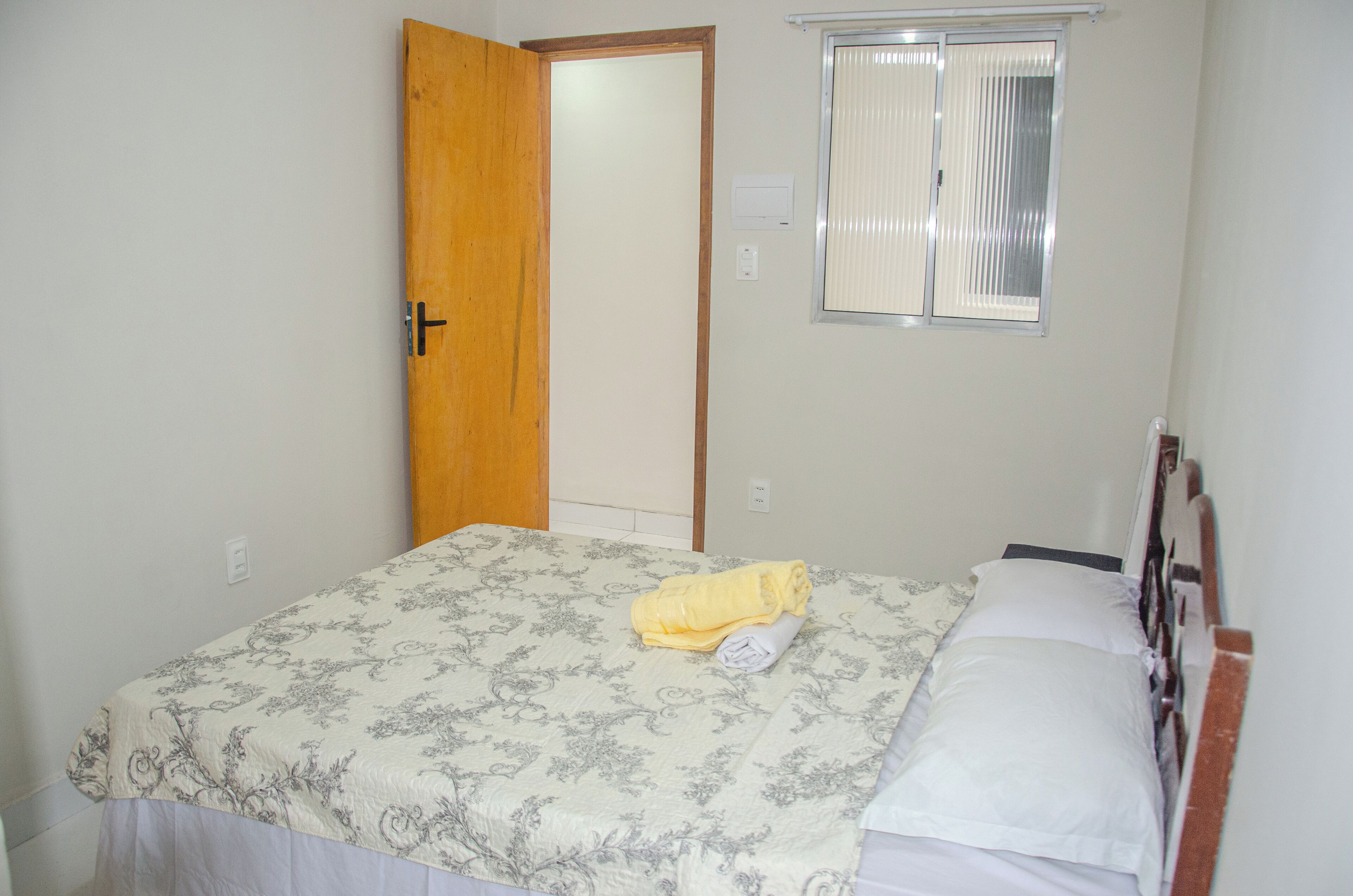 A bedroom with a bed, a window with blinds, a door, and a thermostat on the wall.